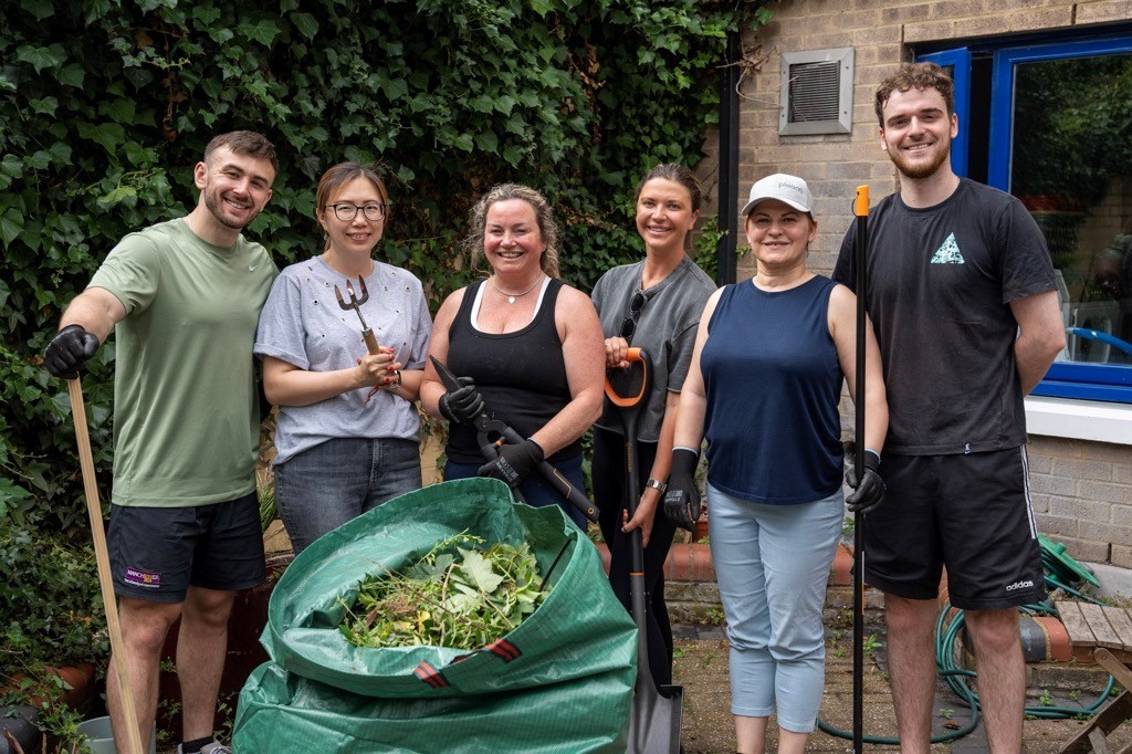 Volunteers from JTI standing in a line holding gardening tools and a bag of garden waste after a volunteering session in the outdoor area of a Look Ahead mental health service.